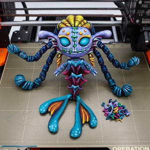 May include: A 3D printed articulated creature figure with a purple head, large teal eyes, and yellow hair. It features long blue and dark blue segmented tentacles, a pink and blue skeletal torso, and a teal tail with yellow fins, plus small scrap pieces.