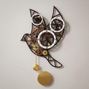 May include: A wall clock shaped like a bird, constructed from dark brown wood with exposed internal gears. It features three white circular dials with black numerals and gold-coloured hands, complemented by a gold pendulum and a circular gold weight.