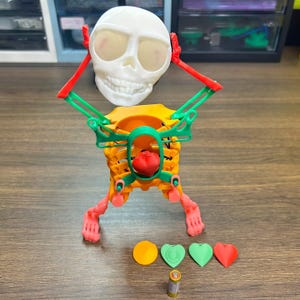 Pode incluir: Uma figura de esqueleto articulada impressa em 3D com crânio branco, caixa torácica laranja, braços verdes e pés cor-de-rosa. Um coração vermelho está dentro da caixa torácica. Quatro pequenas formas planas e uma pilha AA estão à frente.