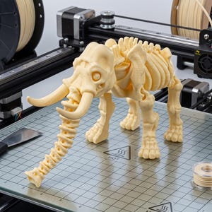 May include: A 3D printed cream-coloured mammoth skeleton figurine with articulated joints, featuring a long trunk, curved tusks, and a detailed rib cage, positioned on the metallic grid print bed of a 3D printer with a visible hot surface warning.