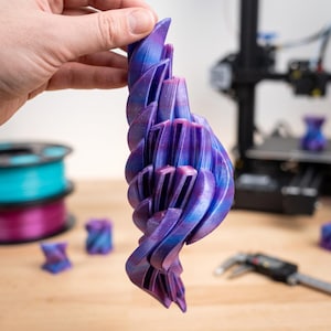 May include: A 3D printed sculpture featuring a vertical, twisting, spiral design with layered segments. The object displays a gradient color pattern shifting between shades of deep purple, vibrant blue, and magenta with a subtle glittery finish.