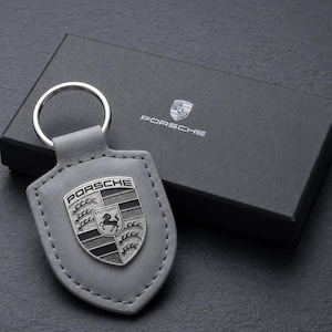 May include: A grey leather keychain featuring a metal shield-shaped emblem with the Porsche crest and text. The keychain is positioned in front of a black rectangular gift box that displays the Porsche logo and text in silver on the lid.