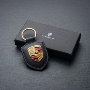 May include: Black leather keychain with a gold ring and a Porsche emblem. The emblem features a gold, red, and black shield with the word "PORSCHE" and a horse. A black box with the Porsche logo is in the background.