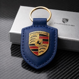 May include: A blue leather keychain with a gold-colored ring. The keychain features the Porsche emblem in gold, red, and black. The keychain is in front of a silver box with the Porsche logo.