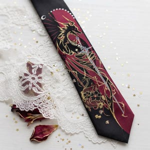 May include: A black necktie featuring a maroon panel with a gold-outlined dragon illustration, a sword, and floral motifs. The design includes a crescent moon and small diamond shapes. The tie is displayed alongside white lace and scattered gold stars.