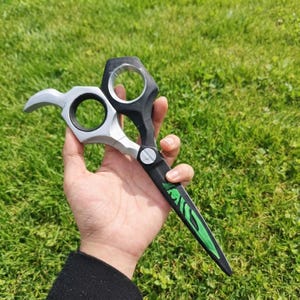 May include: A pair of scissors featuring a silver and black handle with two circular finger holes and a black blade decorated with a bright green graphic design. The tool has a central silver screw and a pointed tip, showcasing a distinct contrast.