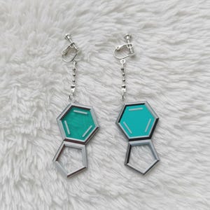 Puede incluir: Un par de pendientes de clip en tono plateado con un diseño geométrico colgante. Cada pendiente consta de un dije hexagonal de color verde azulado con detalles de líneas blancas, unido por una pequeña cadena plateada a un mecanismo de clip metálico.