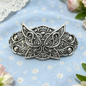 Puede incluir: Una pinza para el cabello de metal plateado con un intrincado patrón en relieve simétrico que se asemeja a alas de mariposa estilizadas. El diseño incluye detalles circulares, líneas onduladas y pequeños motivos florales en toda la superficie.