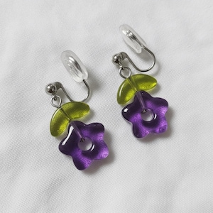 Può includere: Un paio di orecchini a clip con un ciondolo a forma di fiore viola traslucido e un singolo ciondolo a forma di foglia verde lime traslucido. I ciondoli sono fissati a un meccanismo a clip in metallo argentato con un cuscinetto in plastica.