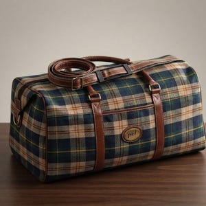 May include: A duffel bag featuring a blue, green, and tan plaid pattern with brown leather trim, handles, and an adjustable shoulder strap. The bag includes a front pocket with a small oval gold-toned metal plaque engraved with the letters pel.