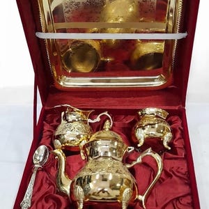 May include: A gold-toned tea set featuring a teapot, a creamer, a sugar bowl, and a spoon, all with intricate embossed patterns. The set is presented in a deep red velvet-lined box, which also includes a matching gold-toned rectangular tray.