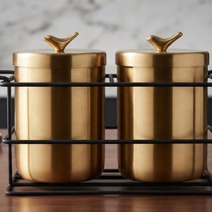 Op de afbeelding: Twee cilindrische goudkleurige metalen bussen met bijpassende deksels, elk voorzien van een kleine vogelvormige handgreep. De bussen worden bij elkaar gehouden in een rechthoekig zwart draadframe met handgrepen aan de zijkanten.
