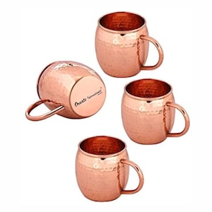 May include: A set of four copper-colored mugs with a hammered texture and curved handles. One mug is positioned on its side, revealing the base with the text Ornate International printed in black. The mugs have a rounded, barrel-like shape.