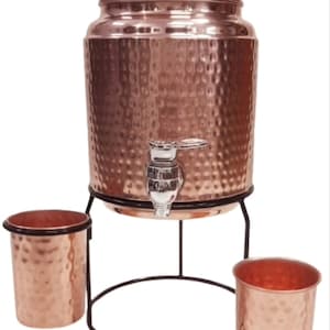 May include: A hammered copper water dispenser with a lid and a silver-toned spigot, resting on a black metal stand. Two matching hammered copper tumblers are positioned on either side of the stand, completing this set of drinkware.