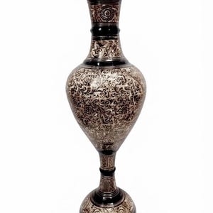 Puede incluir: Un jarrón de metal alto y esbelto con un acabado negro oscuro y grabados intrincados de flores y enredaderas de color claro que cubren el cuerpo bulboso, el cuello acampanado y la base circular, creando un objeto decorativo detallado y contrastado.