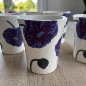 Peut inclure: Un ensemble de tasses en céramique blanche arborant un motif répétitif de grandes fleurs stylisées violettes et bleues avec des tiges vert foncé et de petits bourgeons. Les tasses présentent une forme conique sans anse et un intérieur blanc.