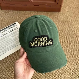 May include: A forest green baseball cap featuring a distressed, frayed brim and a central embroidered patch with a black border and yellow lettering that reads GOOD MORNING. The cap has a structured crown with ventilation eyelets on top.