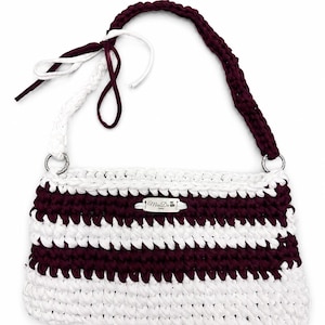 Può includere: Una borsa all'uncinetto fatta a mano con strisce bianche e bordeaux alternate. La borsa presenta un manico bordeaux e lacci bianchi. Anelli in metallo argentato collegano il manico alla borsa. È visibile una piccola targhetta metallica con testo.