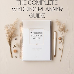 May include: A white wedding planner guide with gold spiral binding, a gold pen, and dried floral accents. The cover reads "WEDDING PLANNER GUIDE" and "THE COMPLETE WEDDING PLANNER GUIDE". The text at the bottom says "Your essential companion for a perfect wedding".