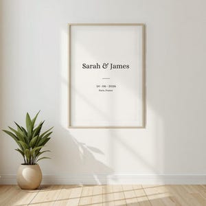 Peut inclure: Une affiche encadrée sur un mur blanc avec le texte "Sarah & James" au-dessus de la date "14.06.2026" et "Paris, France". Une plante en pot est posée sur le sol en bois à gauche du cadre.