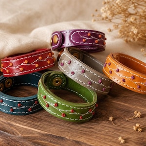 Op de afbeelding: Een collectie van zes leren armbanden in paars, rood, blauwgroen, olijfgroen, grijs en bruin. Elke armband is voorzien van een decoratief wit gestikt rankpatroon met kleine gekleurde knoopjes en een goudkleurige metalen drukknoopsluiting.
