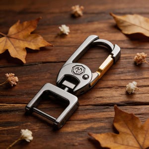 Puede incluir: Un llavero de metal plateado y dorado con un mosquetón y un lazo rectangular. El llavero presenta un emblema circular y está sobre un fondo de madera con hojas de otoño y pequeñas flores.
