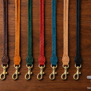 Puede incluir: Siete cordones de cuero finos en marrón, naranja, verde, rojo, azul, beige y negro, cada uno con un mosquetón giratorio de metal dorado en la parte inferior. El texto Tether Tab aparece en la esquina inferior derecha junto a una lengüeta.