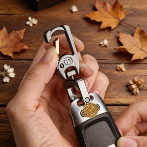 May include: A silver and black key chain with a gold accent. The key chain has a carabiner-style clip and a decorative emblem with a gold and brown design. The key chain is held in a person's hands.