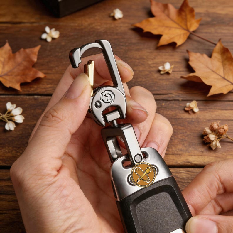 May include: A silver and black key chain with a gold accent. The key chain has a carabiner-style clip and a decorative emblem with a gold and brown design. The key chain is held in a person's hands.