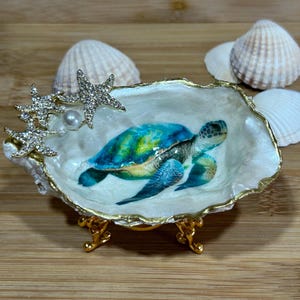 May include: A decorative oyster shell dish featuring a painted sea turtle in shades of blue and green. The shell edge is trimmed in gold, adorned with two rhinestone-encrusted starfish and a single white pearl. It rests on a gold metal stand.