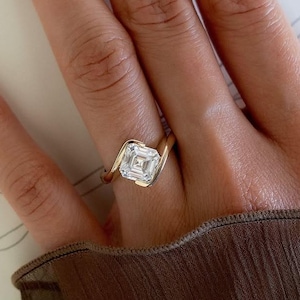 May include: A gold ring featuring a square-cut, clear gemstone set in a bypass-style band. The metal band curves around the central stone, creating a sleek, continuous design that highlights the clarity and geometric shape of the centre stone.