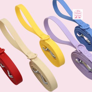Puede incluir: Cuatro correas para perro en rojo, beige, amarillo y lavanda, cada una con un asa de bucle, un mosquetón giratorio de metal y una anilla circular. Un pequeño logotipo cuadrado blanco muestra el texto House and Hounds con un icono de perro.