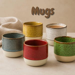 Può includere: Cinque tazze in ceramica con design bicolore, caratterizzate da una parte superiore colorata e maculata e una base color crema. I colori includono blu, giallo, bianco, rosso e verde. La parola Mugs è stampata in marrone sopra gli oggetti.