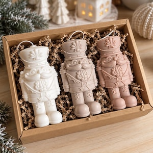 Könnte beinhalten: Ein Set aus drei Nussknacker-Anhängern in einer mit Papierstreifen gefüllten Pappschachtel. Die Figuren sind weiß, hellbeige und altrosa, jede mit einer detaillierten Uniform, einem hohen Hut mit Muschelmotiv und einer Aufhängeschlaufe.