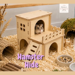 Pode incluir: Um esconderijo para hamster em madeira clara com design de dois andares, apresentando uma pequena entrada em arco, uma varanda com cerca e uma rampa. Um pequeno hamster é visível no nível superior. A estrutura possui janelas recortadas.