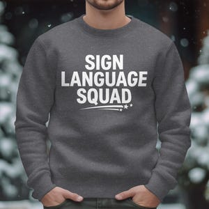 Può includere: Una felpa grigio scuro con la scritta "SIGN LANGUAGE SQUAD" in grandi lettere bianche. Sotto il testo c'è una linea bianca con tre stelle. La felpa è a girocollo.