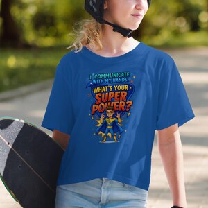 Può includere: Maglietta blu con la scritta "Comunico con le mie mani. Qual è il tuo superpotere?" e una grafica di supereroe. La persona indossa un casco e tiene uno skateboard.