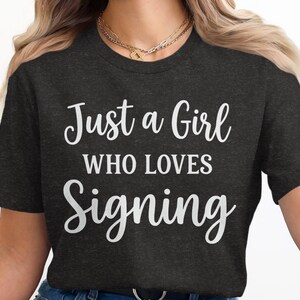 May include: Dark gray t-shirt with the text "Just a Girl Who Loves Signing" in white script. The shirt is a crew neck and appears to be made of a soft material. The overall theme is related to hobbies and interests.
