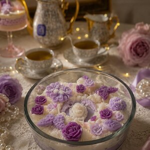 Könnte beinhalten: Dekorative Kerze in einer Glasschale mit floralem Design aus lila und weißen Wachsblumen. Das Bild zeigt Teetassen, eine Teekanne und Perlen, die eine Vintage-Ästhetik erzeugen.