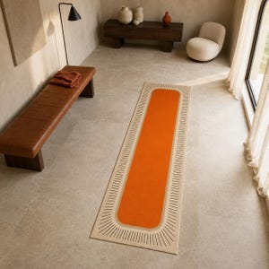 May include: A rectangular runner rug featuring a vibrant orange centre panel surrounded by a beige border with a radiating black line pattern. The rug is positioned on a light-coloured stone floor in a room with a brown leather bench and chair.