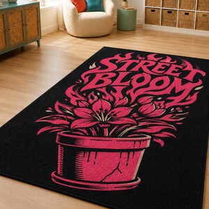 Op de afbeelding: Een rechthoekig zwart vloerkleed met een grafische illustratie van een roze bloem in een pot met vlammen die uit de bloemblaadjes opstijgen. De tekst STREET BLOOM staat in een gestileerd roze lettertype boven de bloem in een gebarsten pot.