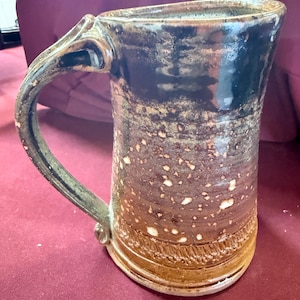 May include: A handcrafted ceramic mug with a large handle. The mug features a dark brown top that fades into a lighter brown with white speckles. The base has a textured, decorative band.