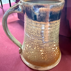 May include: A handmade ceramic mug with a large handle. The mug features a brown and tan speckled glaze with a dark brown band near the rim. The handle is a lighter tan color. The mug has a rustic, handcrafted appearance.