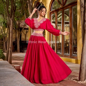 Könnte beinhalten: Ein leuchtend rotes zweiteiliges Set bestehend aus einer langärmeligen bauchfreien Bluse und einem bodenlangen Rock. Die Bluse hat einen V-Ausschnitt und weiße Stickereien entlang des Ausschnitts und der Taille. Der Text Ethnic Wear Special ist sichtbar.