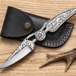 May include: A folding pocket knife with a silver-toned metal handle and blade featuring intricate floral engravings. The knife is positioned over a black leather sheath with a brass-colored snap button closure and a small piece of deer antler.