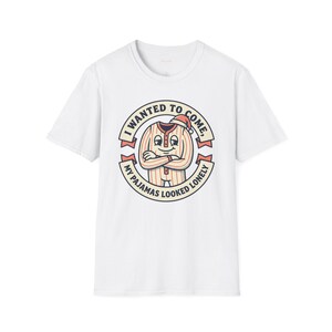 Può includere: T-shirt bianca con un personaggio di pigiama a fumetti con un cappello da Babbo Natale. Il testo recita "I WANTED TO COME, MY PAJAMAS LOOKED LONELY" in un design circolare. Il pigiama è a righe rosse e bianche.