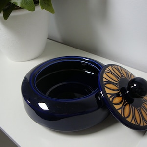 May include: A round, deep, dark blue ceramic bowl with a glossy finish. The matching lid features a central black knob surrounded by a decorative pattern of yellow and orange petals outlined in black, creating a distinct floral-like design.