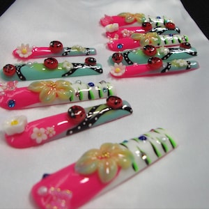 Op de afbeelding: Een set lange, rechthoekige kunstnagels met een levendig ontwerp in felroze, lichtblauwe en witte secties. De nagels zijn versierd met driedimensionale lieveheersbeestjes, kleine witte bloemetjes, groene strepen en kleine kleurrijke strass-steentjes.
