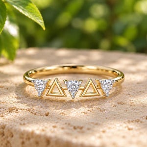 May include: A gold ring featuring a row of five alternating elements: three triangular-cut clear stones and two open gold triangles. The band is smooth and polished gold, showcasing a geometric design with sparkling stones set in metal prongs.