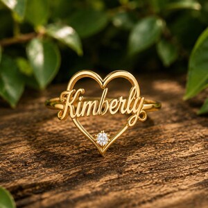 Può includere: Un anello color oro caratterizzato da una montatura a forma di cuore con il nome Kimberly scritto in corsivo al centro. Una singola piccola pietra preziosa rotonda e trasparente è incastonata nella punta inferiore del cuore.